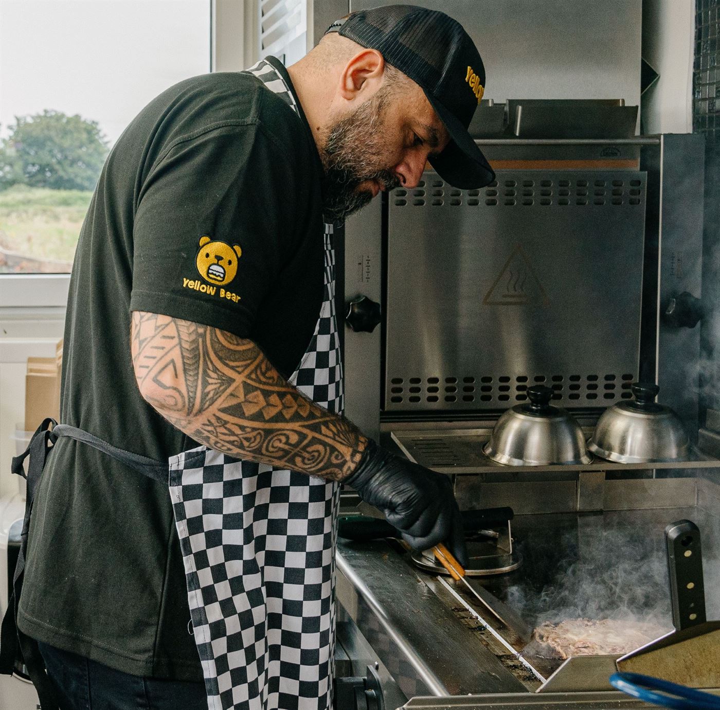 A Yellow Bear chef in a branded cap and chequered apron smashing a burger patty on the griddle